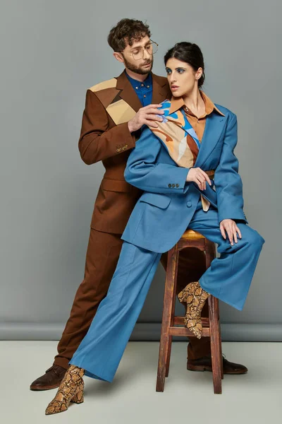 Брюнетка жінка сидить на дерев'яному табуреті біля стильного чоловіка в окулярах, моделі в костюмах, сірий фон — стокове фото