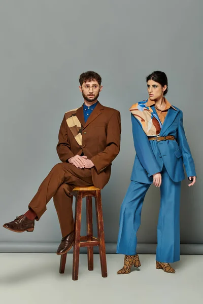 Мужчина в очках сидит на деревянном стуле, женщина позирует на сером фоне, модная пара в костюмах — стоковое фото