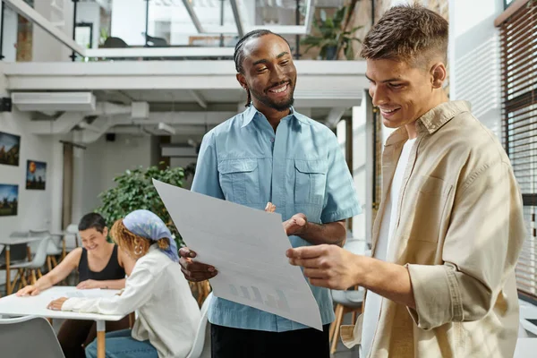 Cheerful interracial men looking at project on paper, office workers smiling near female colleagues — Stock Photo