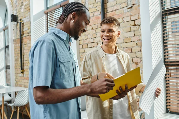 Happy man looking at african american colleague, startup project, holding paper and notebook — Stock Photo