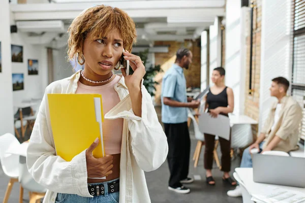 Donna afroamericana preoccupata con bretelle che parlano su smartphone, cartella, startup, gen z, coworking — Foto stock