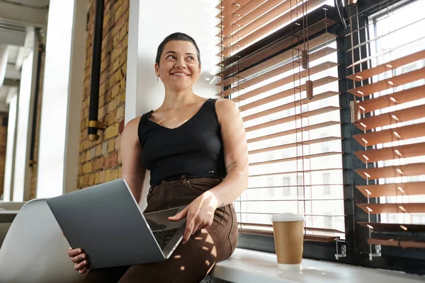 Mujer tatuada alegre usando el ordenador portátil, sentado en el alféizar de la ventana, café para llevar, proyecto de la puesta en marcha, coworking - foto de stock