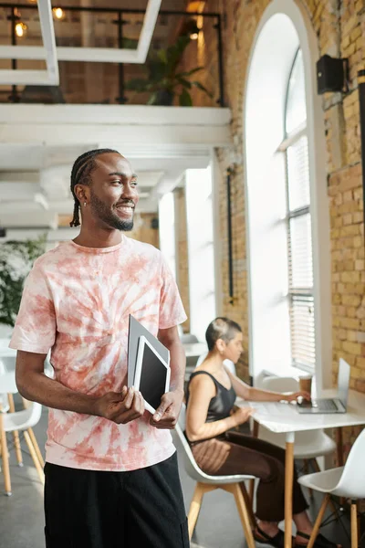 Homem americano africano alegre segurando tablet, pasta, olhando para longe perto de colega de trabalho feminino, inicialização — Fotografia de Stock