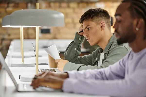 Homem pensativo verificando documentos, trabalhando no escritório, colegas diversos, co-working, laptops, startup — Fotografia de Stock