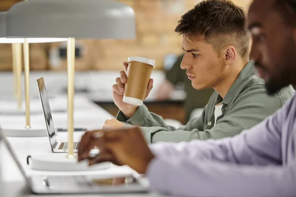 Joven que sostiene el café para ir y el uso de la computadora portátil cerca de un colega afroamericano, startup, gen z - foto de stock