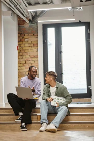 Afrikanisch-amerikanischer Mann zeigt männlichen Kollegen Projekt auf Laptop, sitzt auf Treppe und teilt Ideen — Stockfoto
