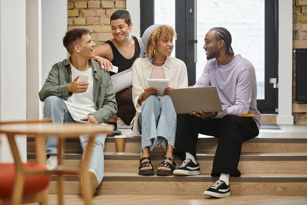 Cheerful diverse team holding gadgets and discussing project, sitting on stairs in modern coworking — Stock Photo