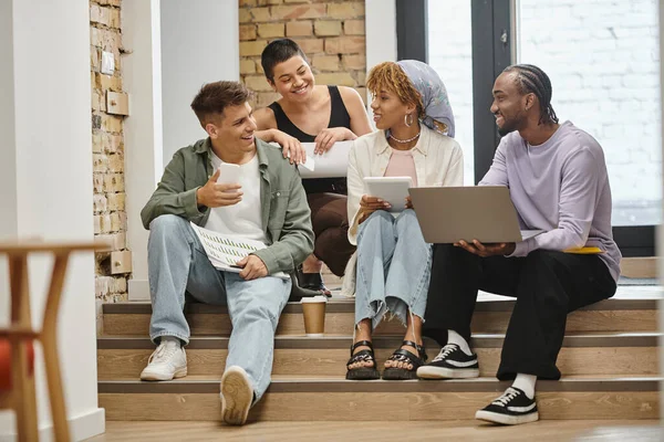 Positive diverse team holding gadgets and discussing project, sitting on stairs in modern coworking — Stock Photo