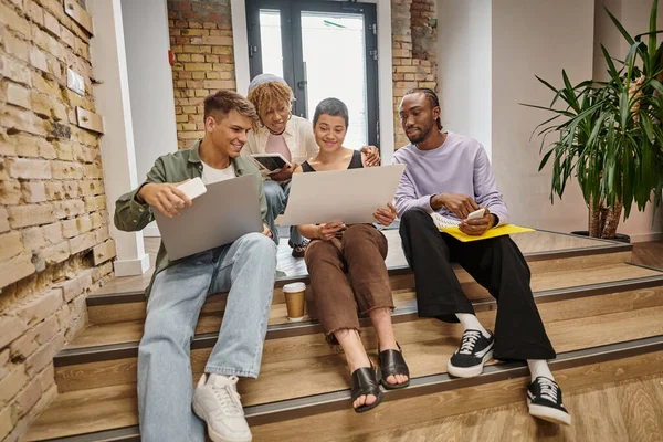Equipe interracial feliz olhando para laptop, segurando gadgets, sentado em escadas em co-working, inicialização — Fotografia de Stock