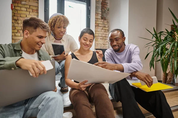 Happy interracial team holding gadgets, sitting on stairs in coworking, looking at startup project — Stock Photo