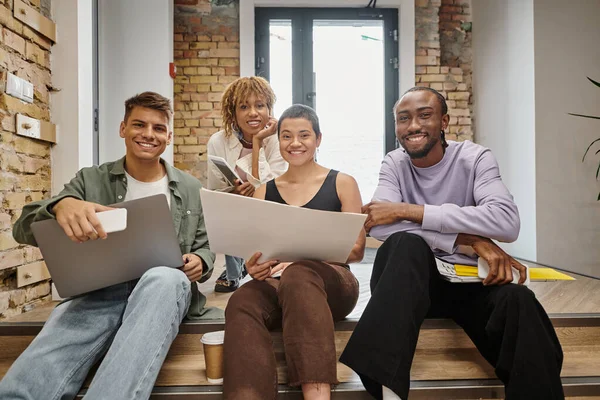 Startup, happy interracial team looking at camera, holding gadgets, sitting on stairs in coworking — Stock Photo