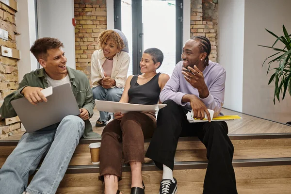 Startup, happy diverse team chatting, holding gadgets, sitting on stairs in modern coworking, tech — Stock Photo