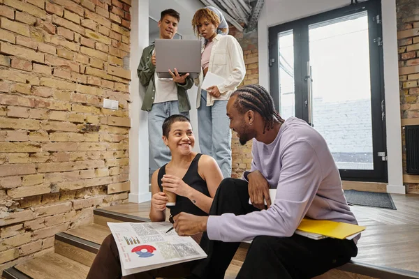 Gente de negocios diversa feliz discutiendo gráficos, sentado en escaleras, puesta en marcha, coworking moderno - foto de stock