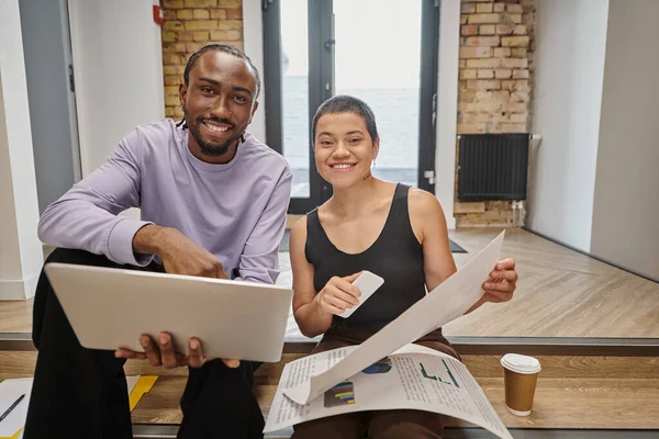 Happy man and woman holding gadgets and graphs, interracial colleagues looking at camera, startup — Stock Photo