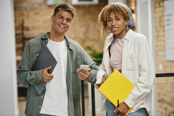 Hombre joven mostrando teléfono inteligente a la mujer afroamericana feliz en aparatos ortopédicos, coworking, startup, trabajo en equipo - foto de stock