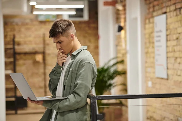 Homem pensativo usando laptop, de pé em co-trabalho moderno, ambiente de escritório, geração z, inicialização — Fotografia de Stock