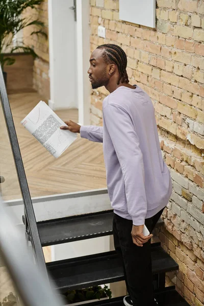 African american man holding smartphone and graphs, walking on stairs in coworking, gen z, startup — Stock Photo