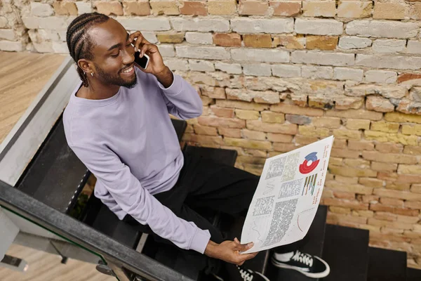 Allegro uomo afroamericano che tiene grafici, seduto sulle scale in coworking, parlando su smartphone — Foto stock