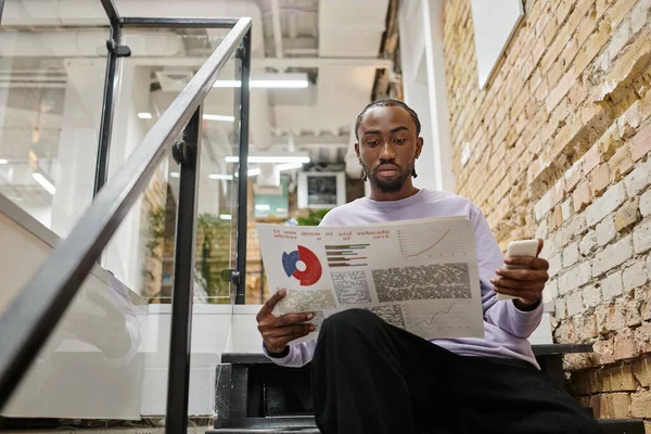 Geschäft, afrikanisch-amerikanischer Mann hält Graphen und Smartphone, sitzt auf Treppen, Coworking, Start-up — Stockfoto