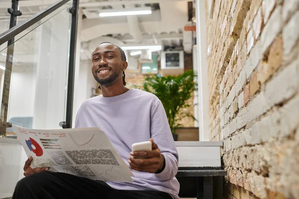 Positive african american man holding graphs and smartphone, sitting on stairs, coworking, startup — Stock Photo