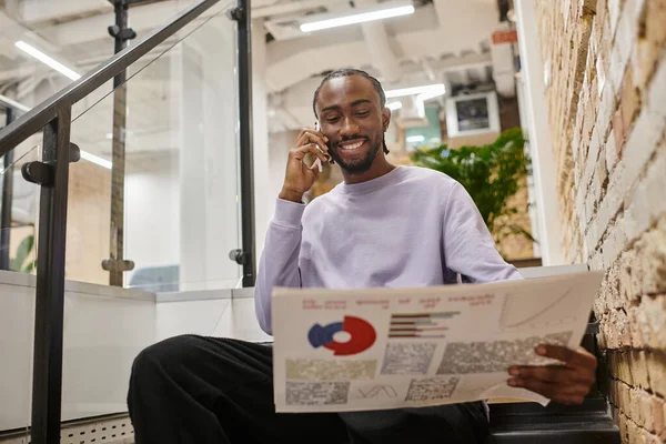 Homem afro-americano positivo segurando gráficos e falando no smartphone, sentado em escadas, inicialização — Fotografia de Stock