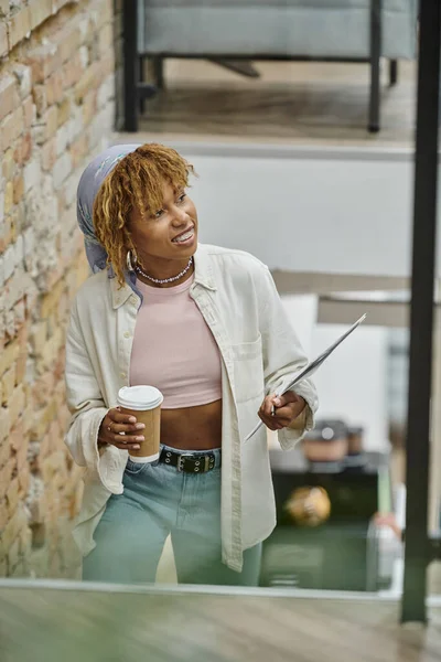 Heureuse femme afro-américaine avec bretelles tenant tasse en papier, café et documents, projet de démarrage — Photo de stock