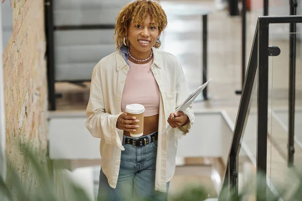 Femme afro-américaine positive avec bretelles tenant tasse en papier, café et documents, projet de démarrage — Photo de stock