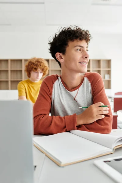Sonriente adolescente colegial sosteniendo lápiz cerca de cuaderno y dispositivos durante la lección en la escuela - foto de stock