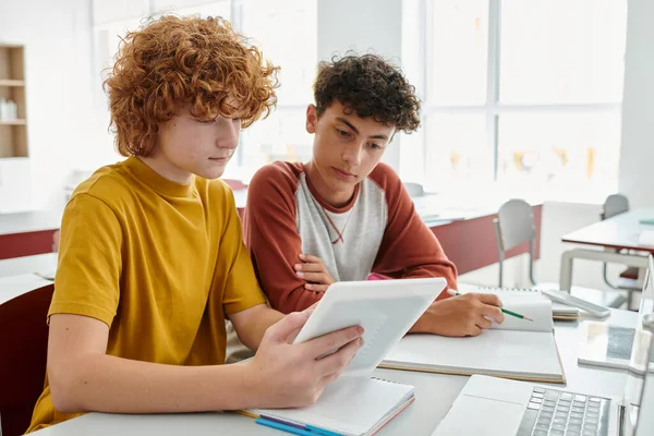 Teenage schoolboys using digital tablet together near notebooks on desk during lesson in classroom — Stock Photo