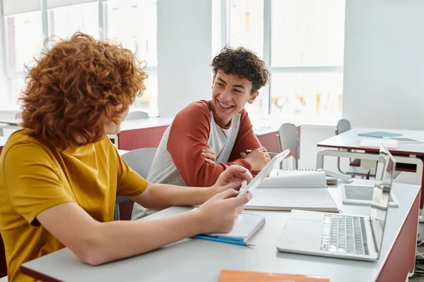Fröhlicher Schüler spricht mit Freund, während er digitales Tablet in der Nähe von Notebooks auf dem Schreibtisch im Klassenzimmer benutzt — Stockfoto