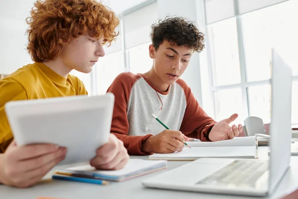 Teenage schoolboy writing on notebook near redhead friend with digital tablet in classroom — Stock Photo