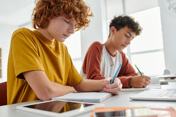 Redhead schoolboy writing on notebook near devices and blurred classmate in school at background — Stock Photo