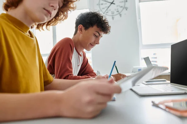 Teenage schoolboy writing while sitting near classmate and devices in classroom at background — Stock Photo