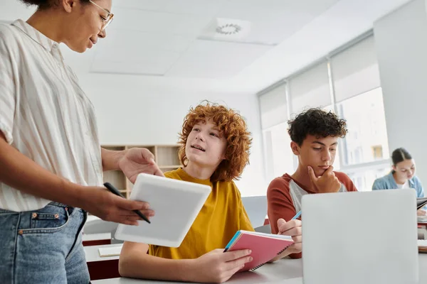 Teen schoolboy talking to african american teacher during lesson with devices in classroom — Stock Photo