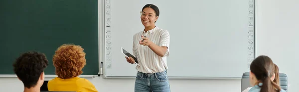 Insegnante afroamericano sorridente con dispositivo che indica gli alunni durante la lezione in classe, striscione — Foto stock