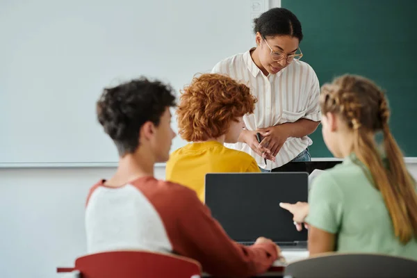 African american teacher standing near blurred pupils with laptops during lesson in classroom — Stock Photo