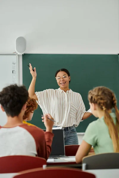 Profesora afroamericana positiva hablando con alumnos adolescentes borrosos cerca de la computadora portátil en el aula en la escuela - foto de stock