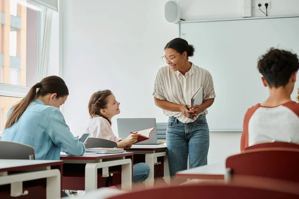 Adolescente sonriente colegiala sosteniendo cuaderno cerca de profesor afroamericano y compañeros de escuela en el aula - foto de stock