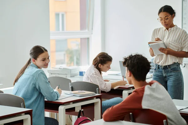 Smiling schoolgirl looking at classmate near devices and african american teacher in classroom — Stock Photo