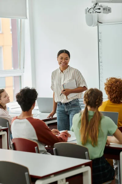 Молодий усміхнений афроамериканський вчитель з ноутбуком розмовляє зі школярами під час уроку на заняттях — стокове фото