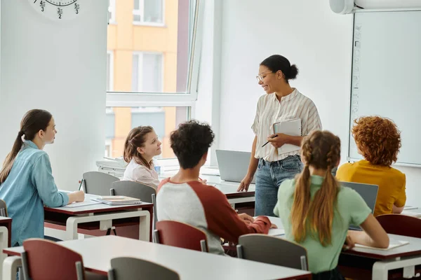 Smiling schoolgirl looking at african american teacher with notebook during lesson in classroom — Stock Photo
