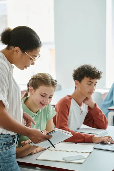 African american teacher holding notebook near positive schoolgirl during lesson in classroom — Stock Photo