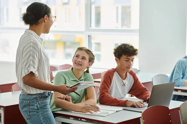 Smiling schoolgirl looking at african american teacher with notebook near classmates in classroom — Stock Photo