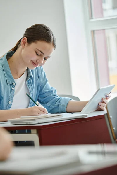 Smiling teenage pupil using digital tablet and writing on notebook during lesson in classroom — Stock Photo