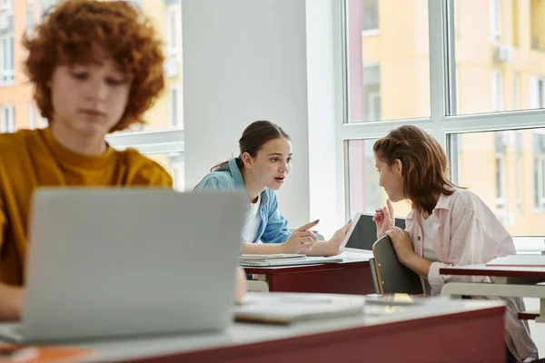Teenage classmates talking and pointing with fingers near devices and notebook in classroom — Stock Photo