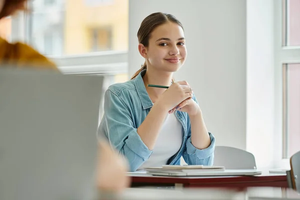 Smiling teenage schoolgirl looking at camera and holding pen near devices during lesson in classroom — Stock Photo