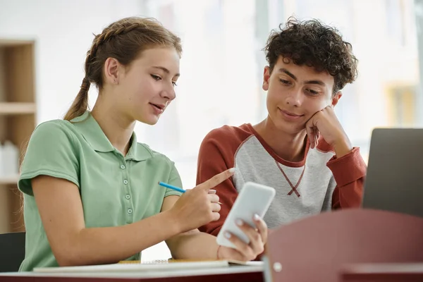 Усміхнена школярка-підліток вказує пальцем і тримає смартфон біля однокласника в класі — стокове фото