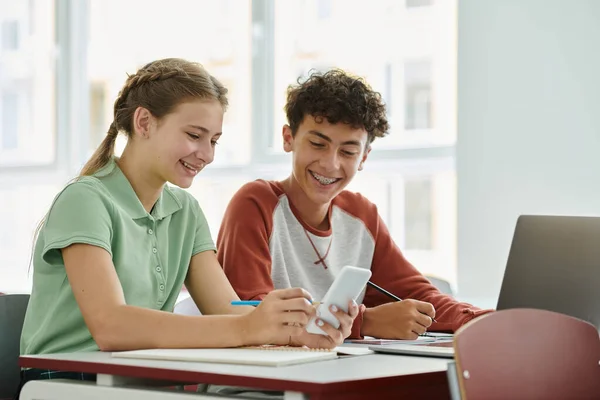 Sorrindo colegas adolescentes usando smartphone juntos perto de dispositivos em sala de aula na escola — Fotografia de Stock