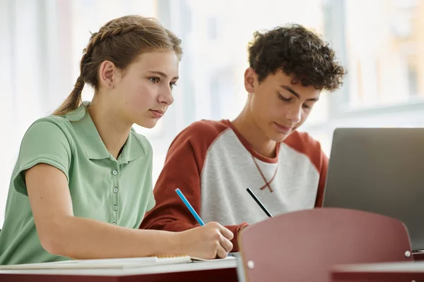 Teenage schoolgirl writing on notebook near blurred classmate and laptop during lesson in class — Stock Photo