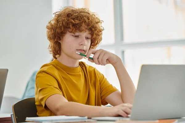 Rossa adolescente scolaro in possesso di matita e utilizzando il computer portatile durante la lezione in aula in background — Foto stock